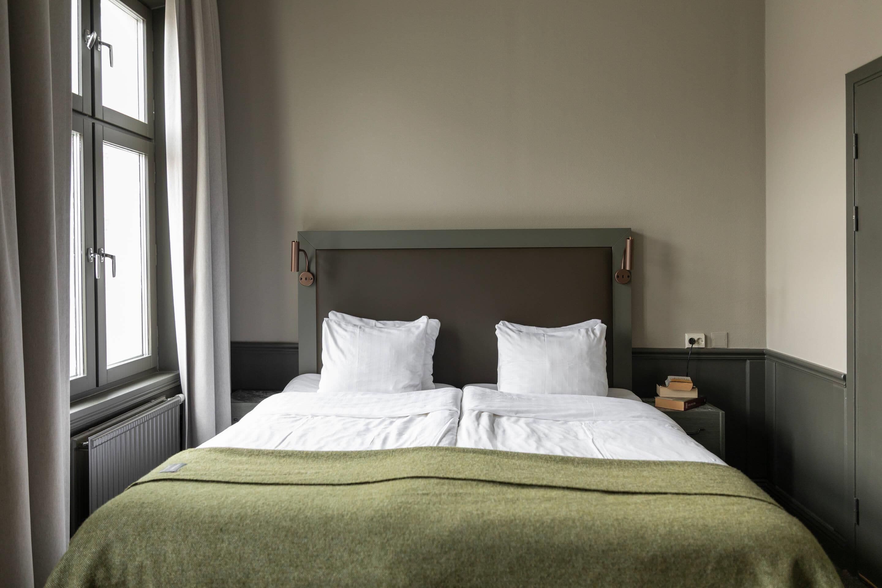 A neatly made bed with white linens and a green throw blanket, a nightstand with a book and a lamp, and a window with curtains.