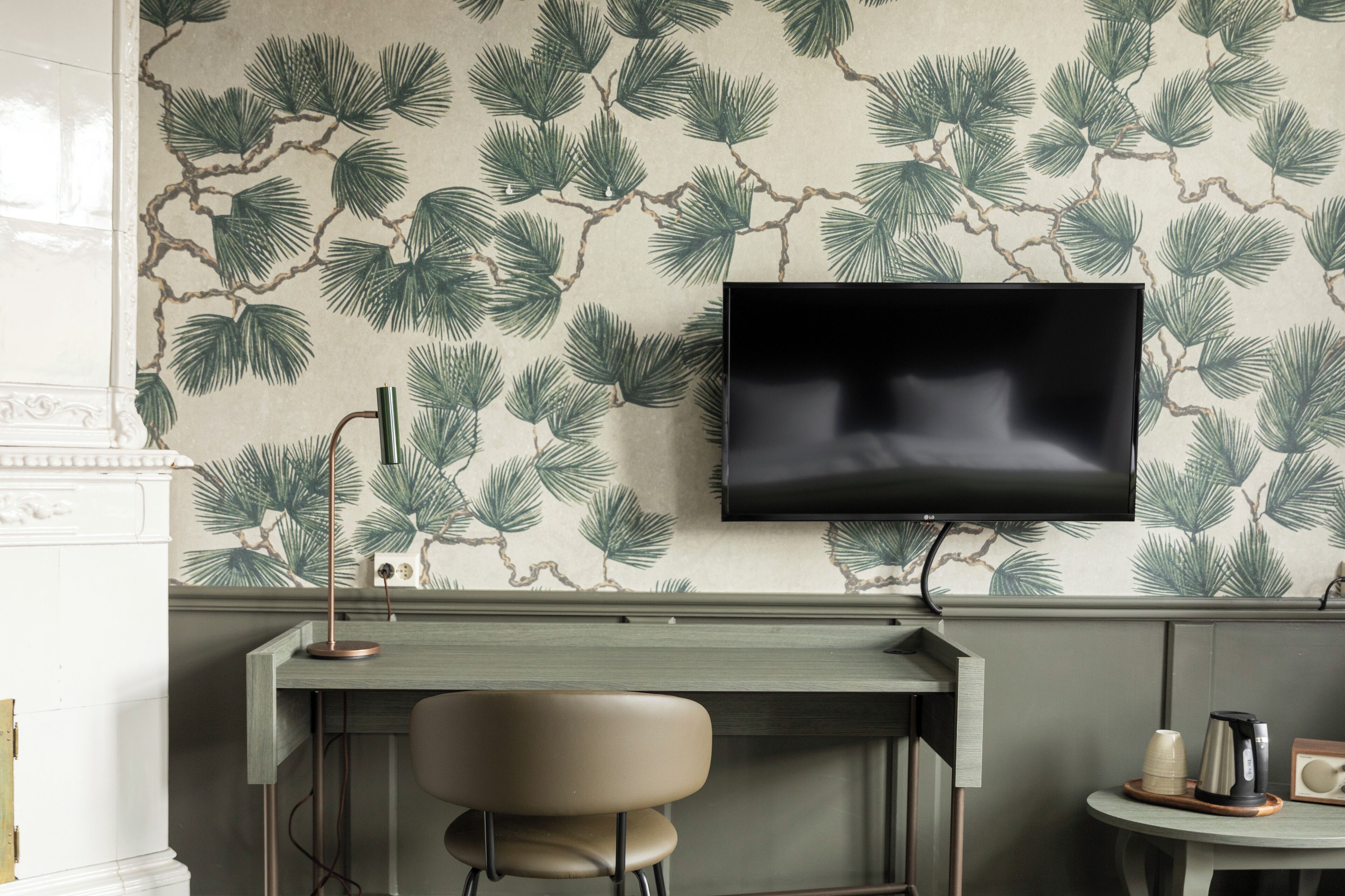 Una habitación con televisión montada en la pared, un escritorio con una silla y una mesita con cafetera y tazas.