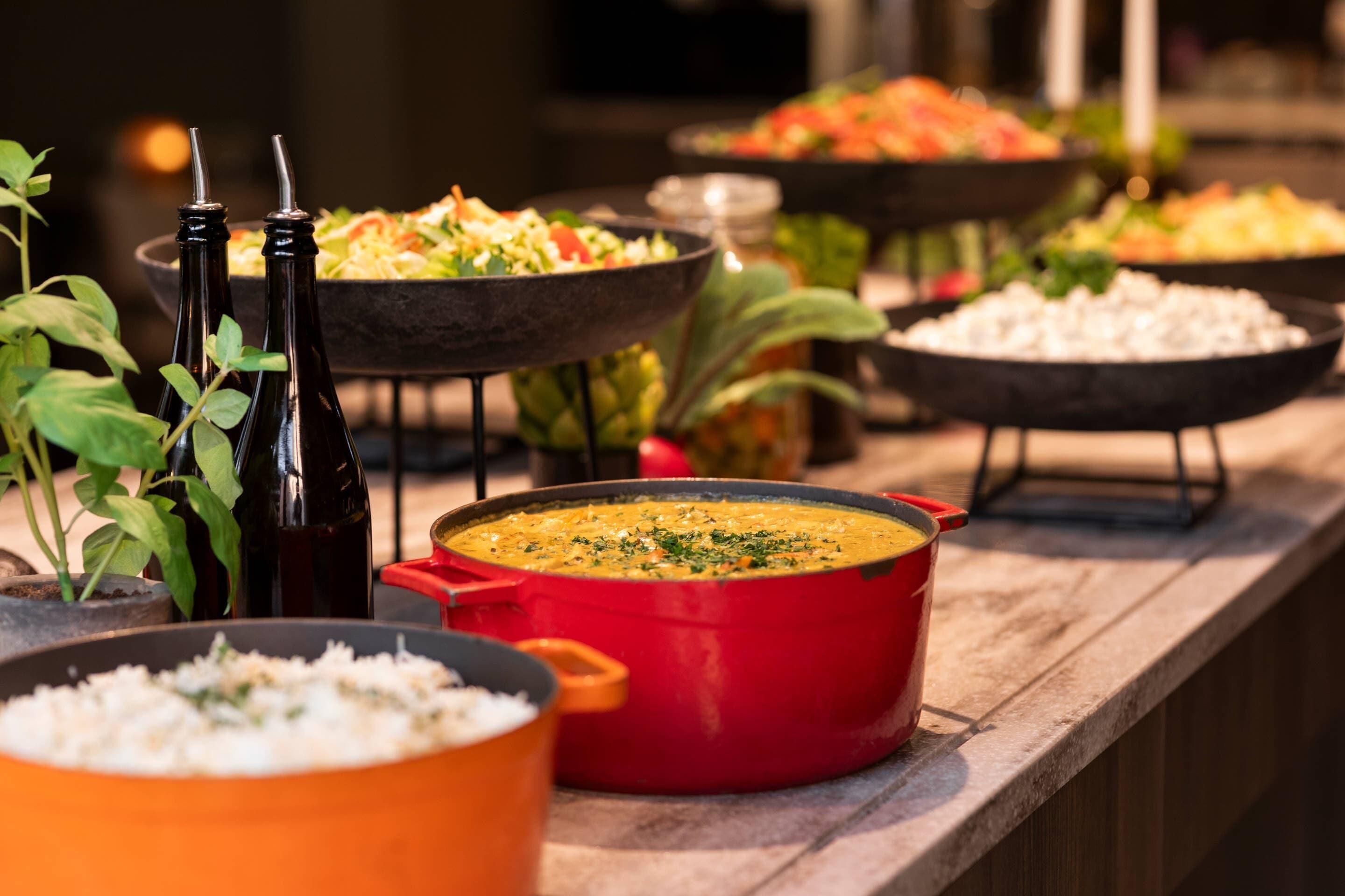 Una mesa de buffet con diversos platos, incluyendo una olla roja de sopa, tazones de arroz y botellas de salsa.