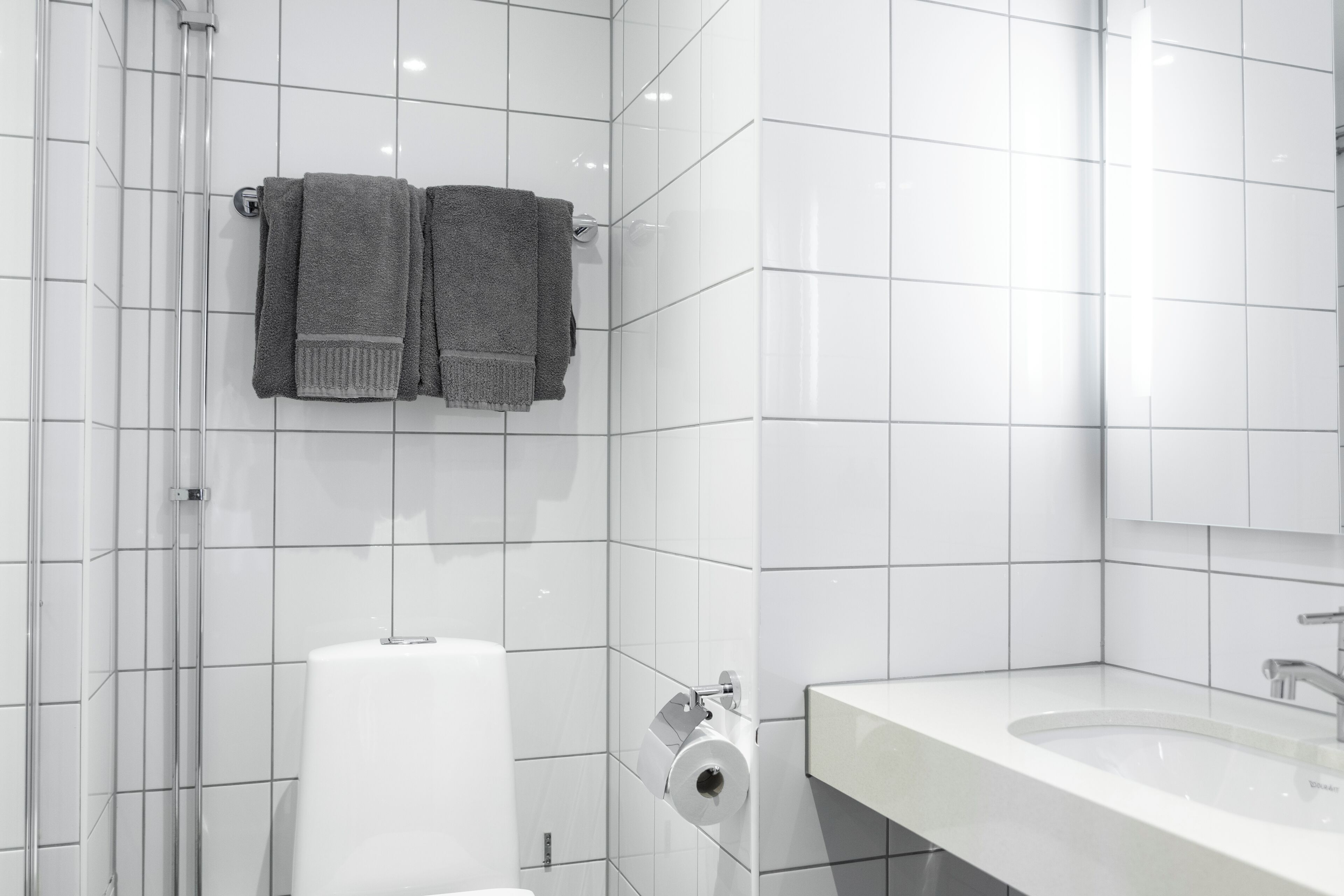 Un baño moderno con azulejos blancos, un inodoro, un lavamanos y dos toallas colgadas en un perchero.