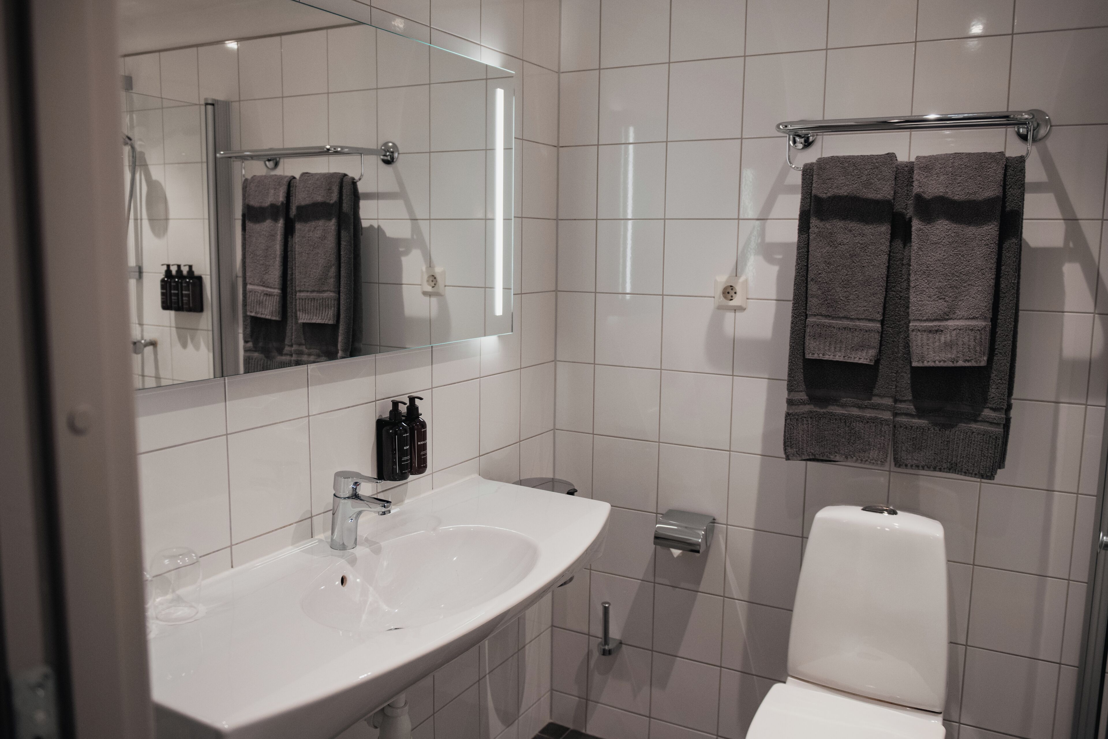 Un baño moderno con un lavamanos blanco, un espejo y dos toallas colgadas en un perchero.