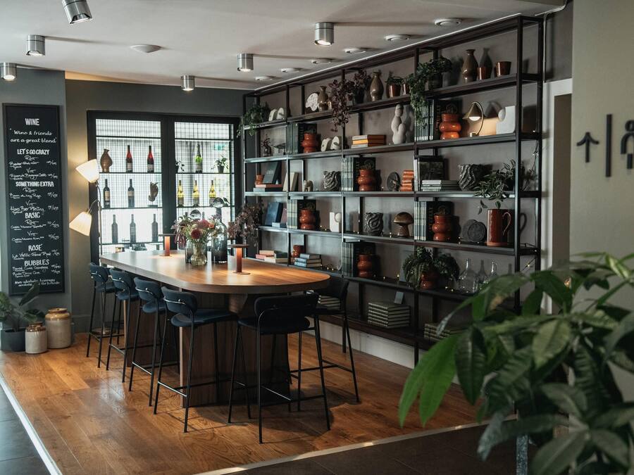 Una zona de bar moderna con mostrador de madera, taburetes negros y estanterías con varias botellas y objetos decorativos.