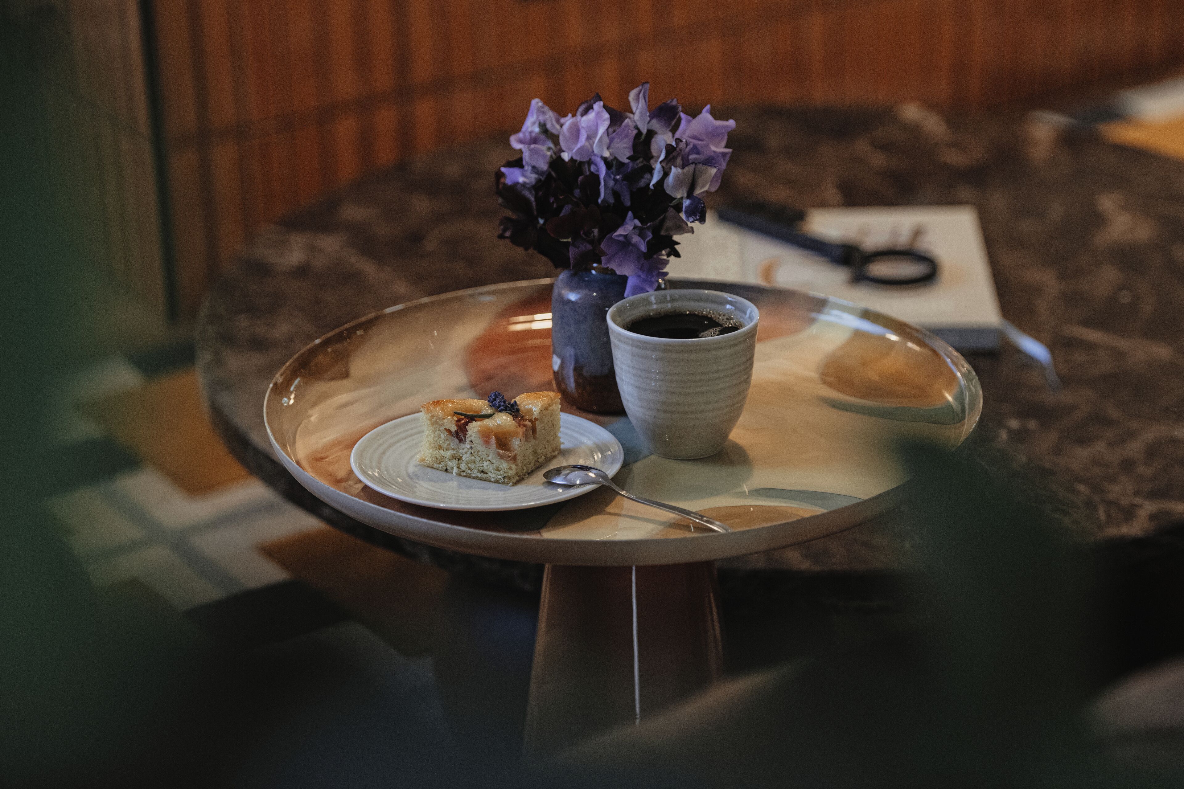 A table with a glass top, a vase with purple flowers, a cup of coffee, a spoon, and a slice of cake on a plate.