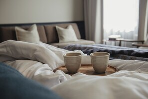 Un lit bien fait, avec deux tasses en céramique posées sur un plateau en bois, une tête de lit et une fenêtre avec des rideaux.