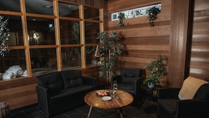 A cozy lounge area with a wooden floor, a round wooden table, a black sofa, and two armchairs. There are potted plants on the walls and a large window with a view of the outside. - Home Hotel Kompaniet (Nykoping)