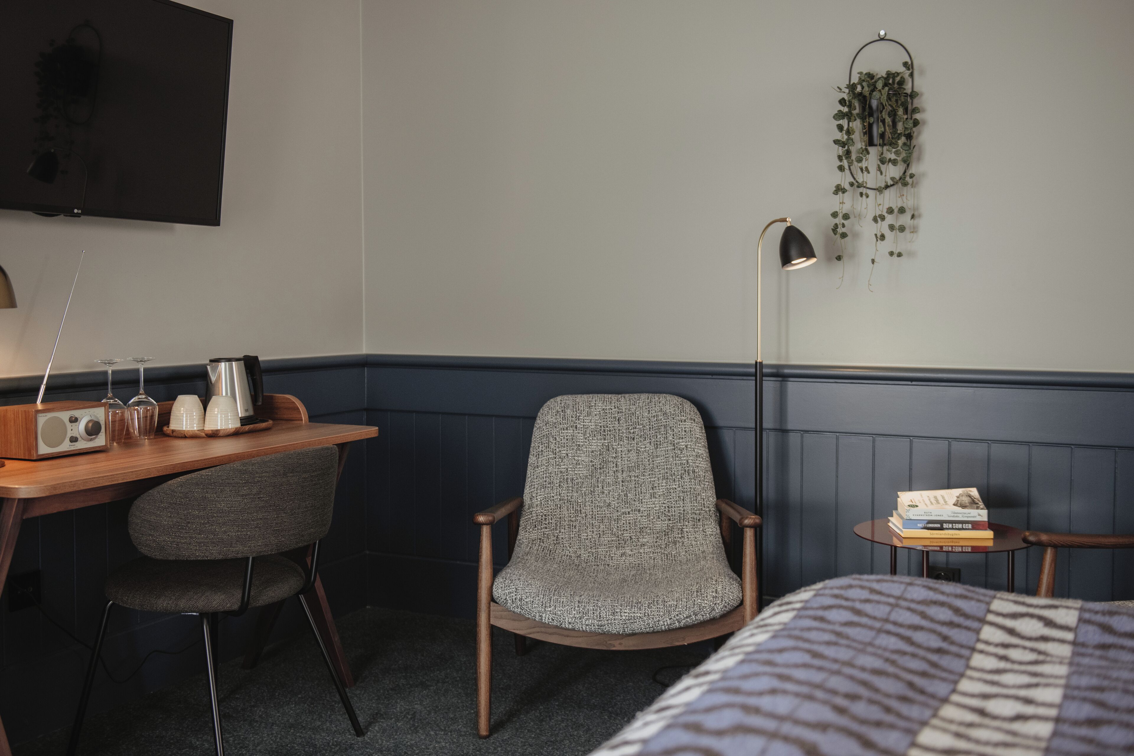 Habitación de hotel con un escritorio de madera, dos sillas, una cama con colcha estampada, un televisor de pantalla plana montado en la pared, una lámpara de pie y una mesita con libros y una planta.