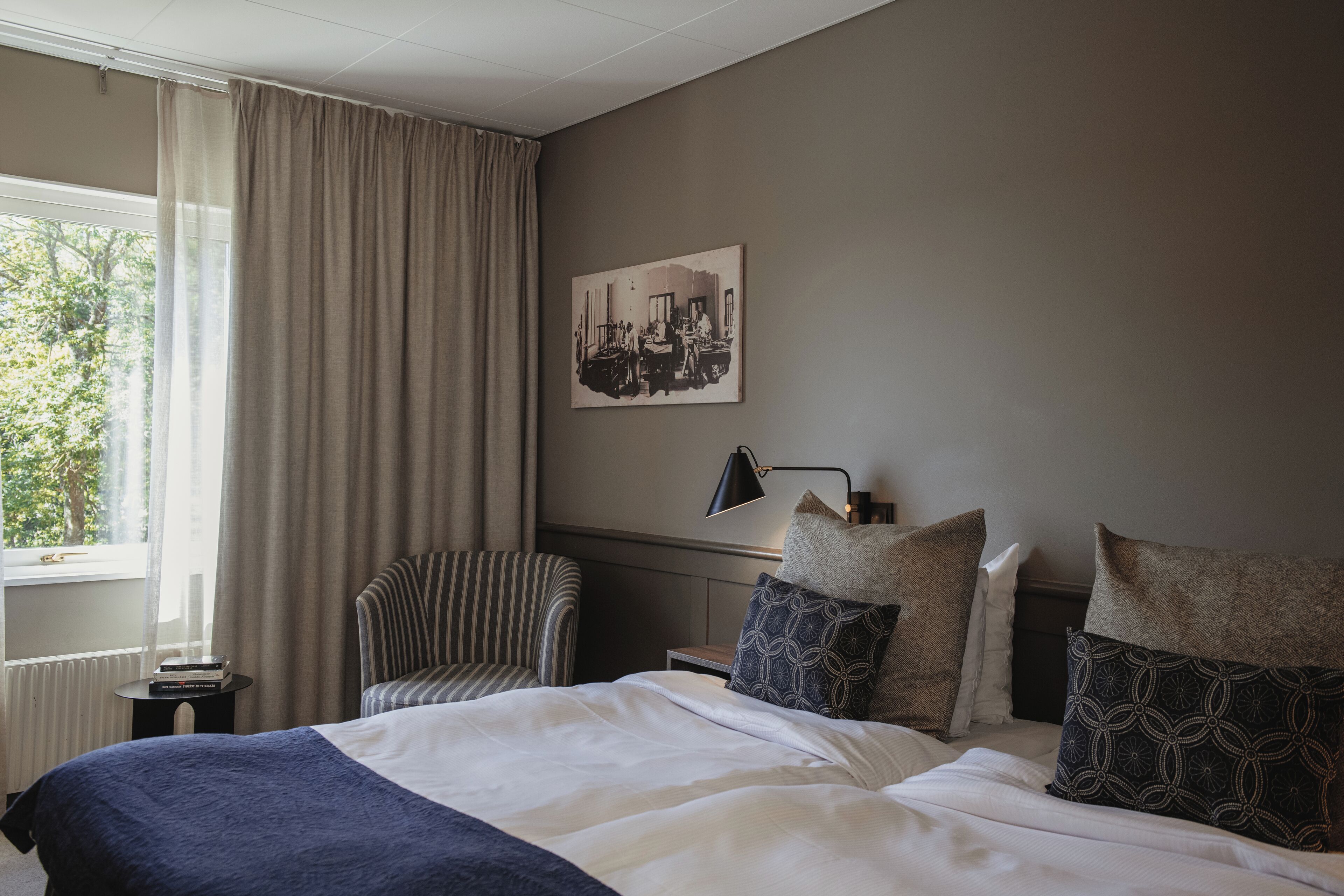 A hotel room with a large bed, a chair, a bedside table, a lamp, and a window with curtains.
