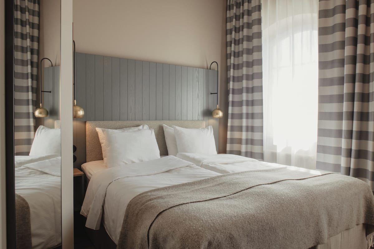 Habitación de hotel con cama, cabecero, lámparas de noche y una ventana con cortinas.