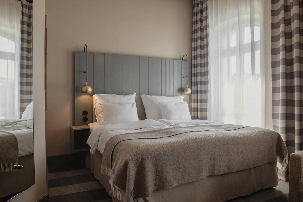 Una habitación de hotel con una cama, lámparas de noche, cabecero y una ventana con cortinas.