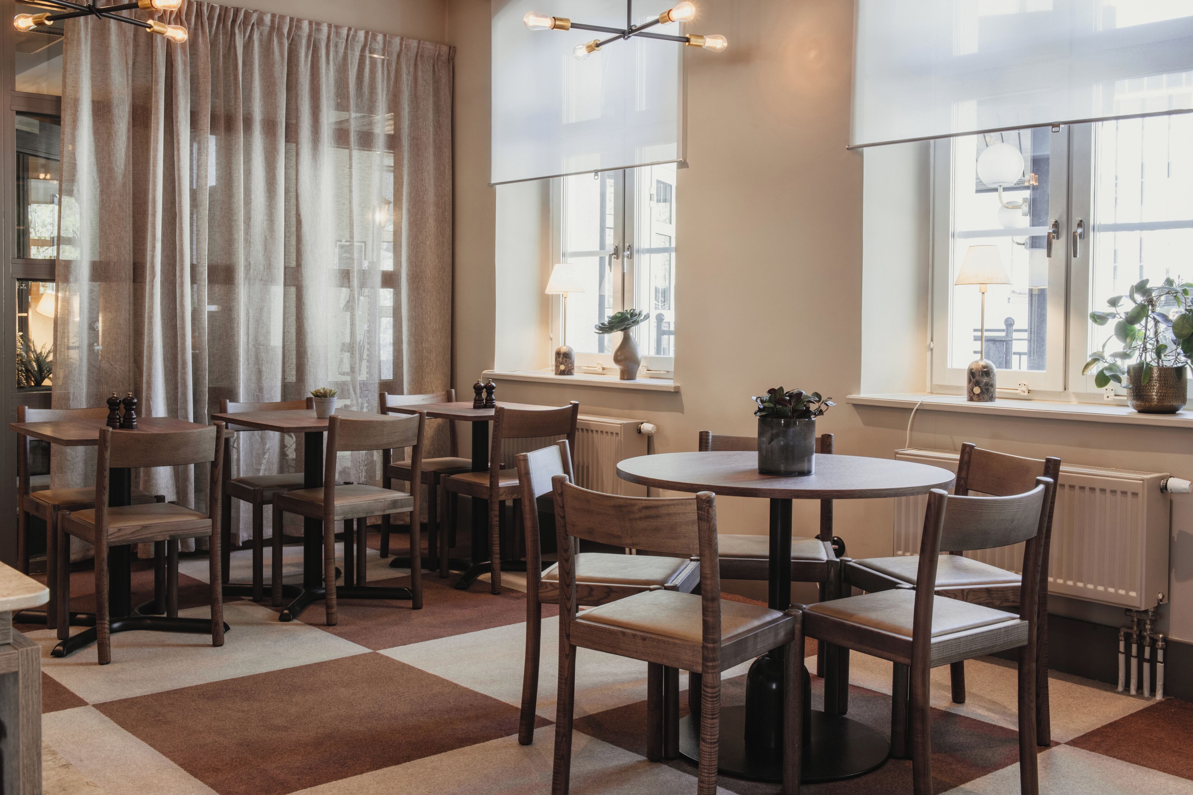A dining area with tables and chairs, large windows with curtains, and a radiator.