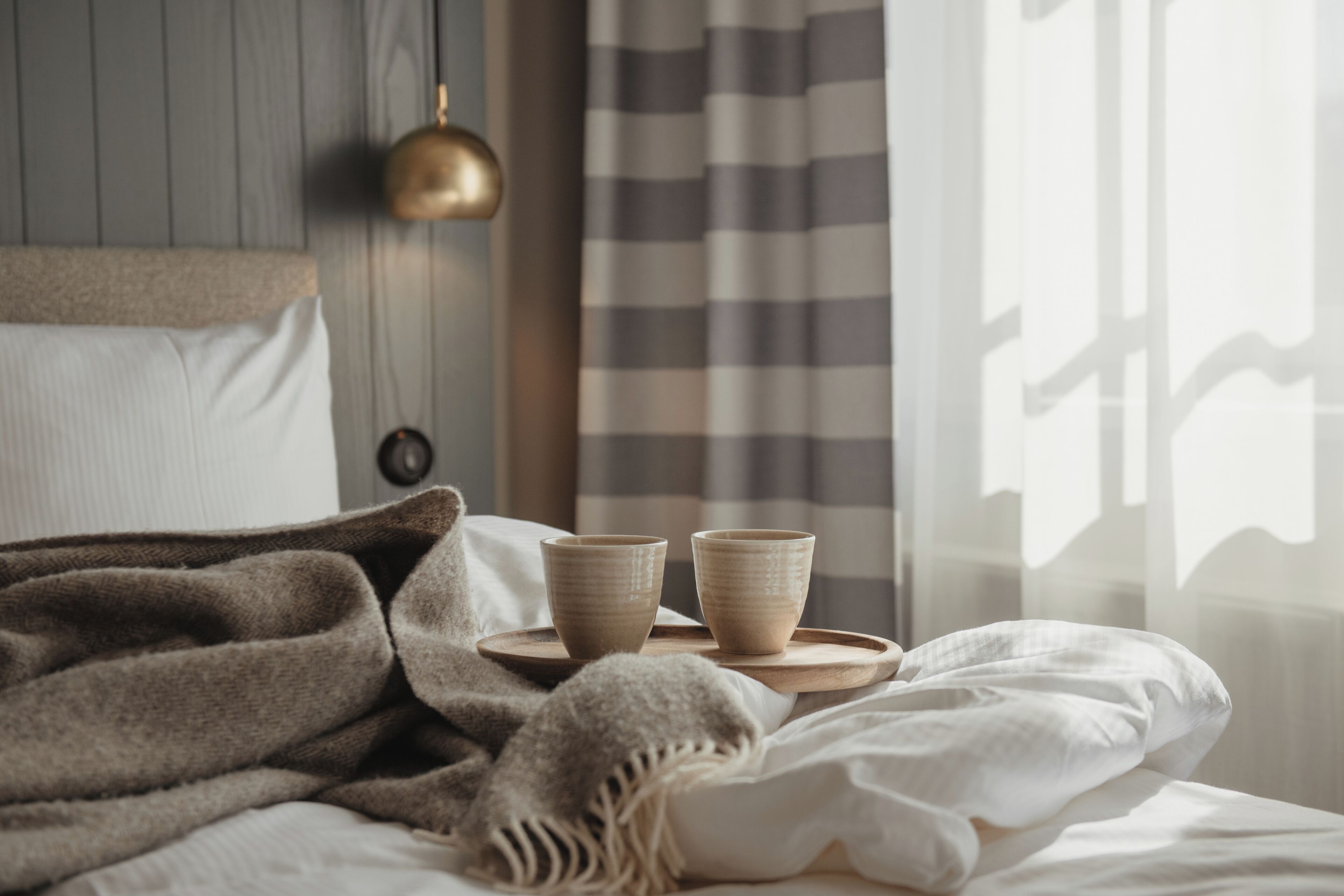 A bed with a blanket, two cups on a tray, a lamp, and a window with curtains.