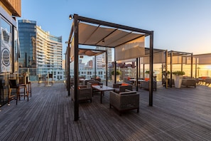 En tagterrasse med udendørs møbler, et pergola-lignende stillads og udsigt over bybilledet.