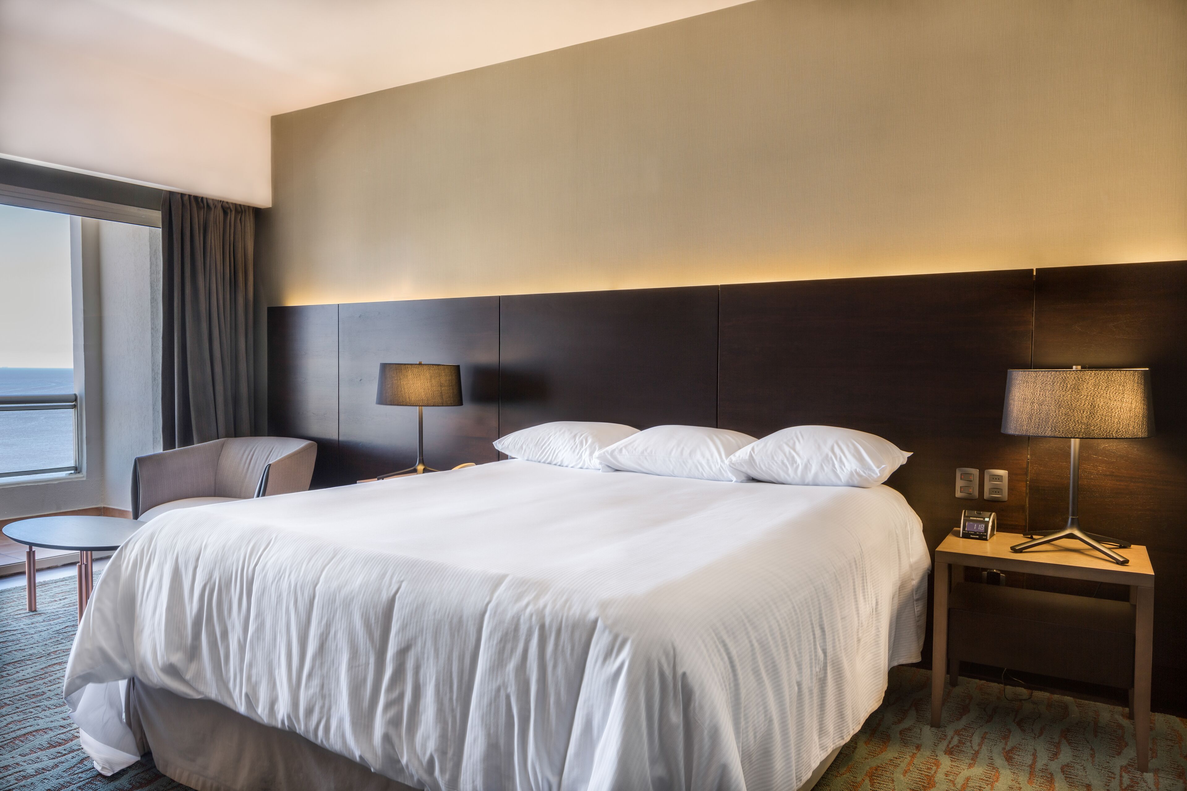 Habitación de hotel con una cama grande, una mesita de noche con lámpara, una silla y vista al mar a través de una ventana.