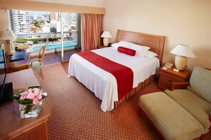 A hotel room with a large bed, two bedside tables, a desk, a chair, and a view of a pool and buildings.