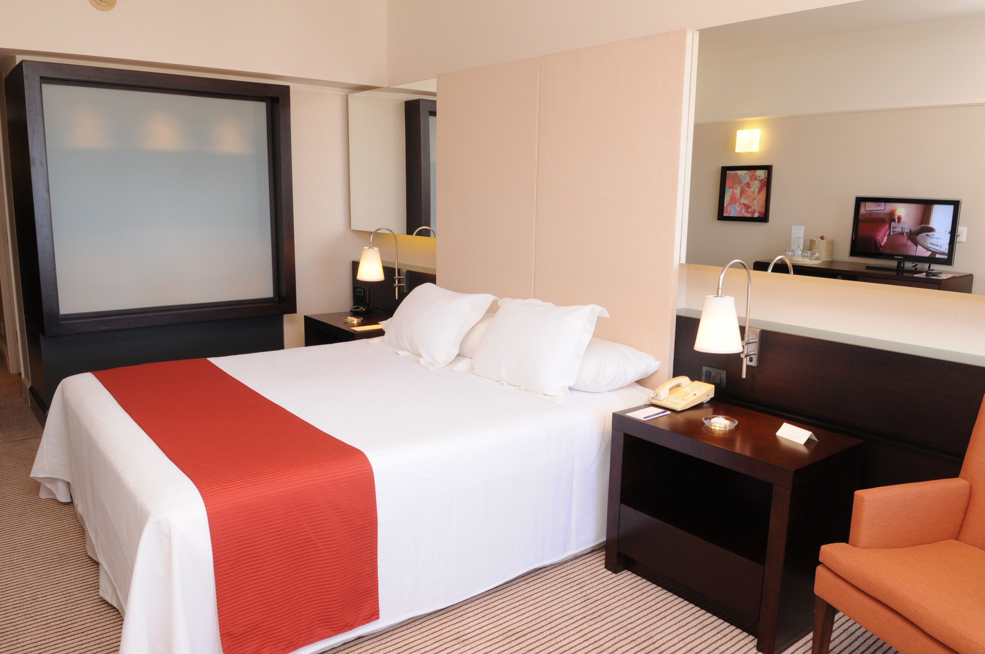 A hotel room with a large bed, a bedside table with a lamp, a chair, a TV, and a mirror.