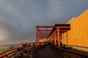 A wooden boardwalk leading to a beachside restaurant with outdoor seating and string lights.