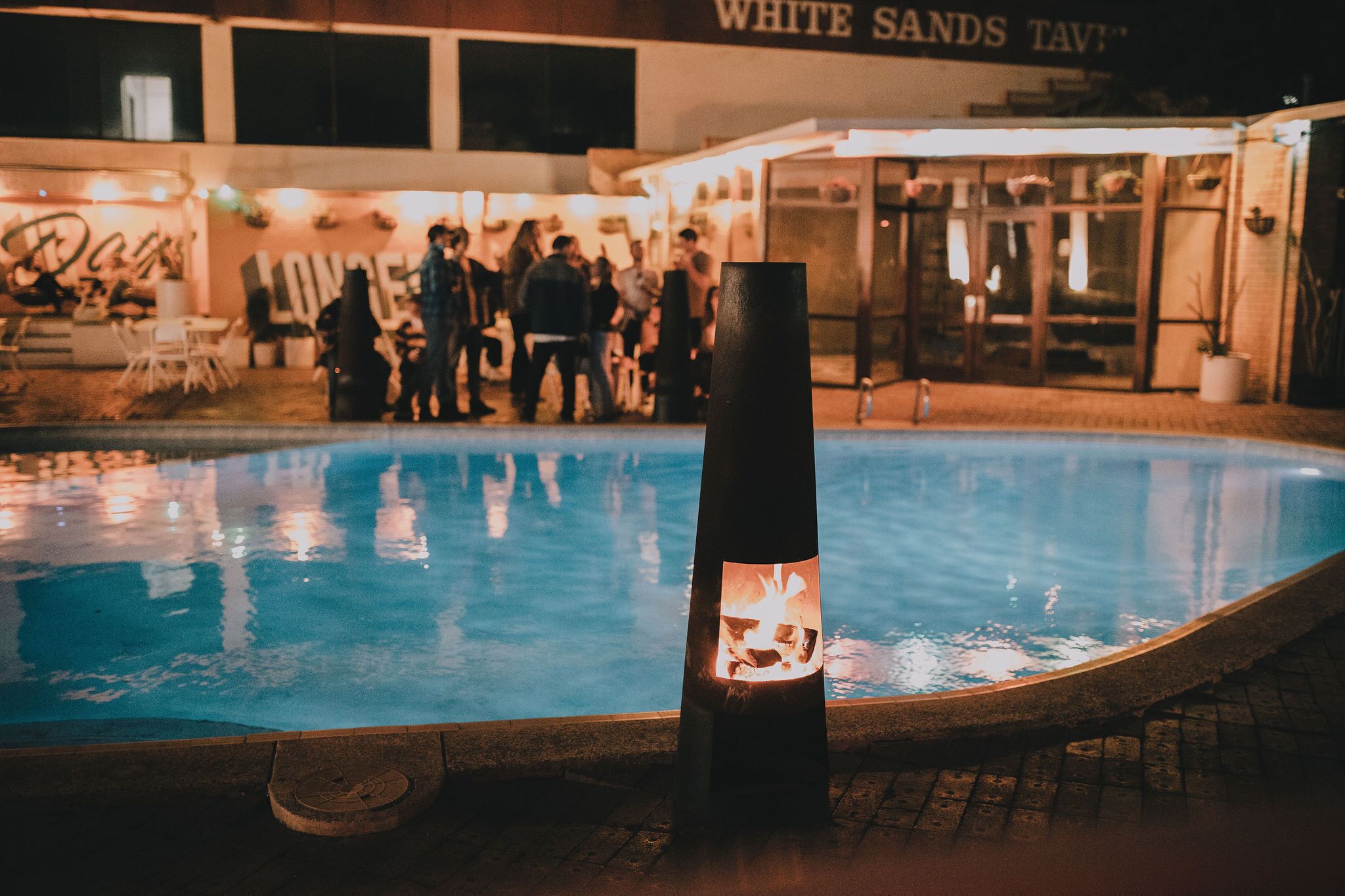 Una zona de piscina con un elemento decorativo de fuego al frente y un edificio con un letrero en el fondo.