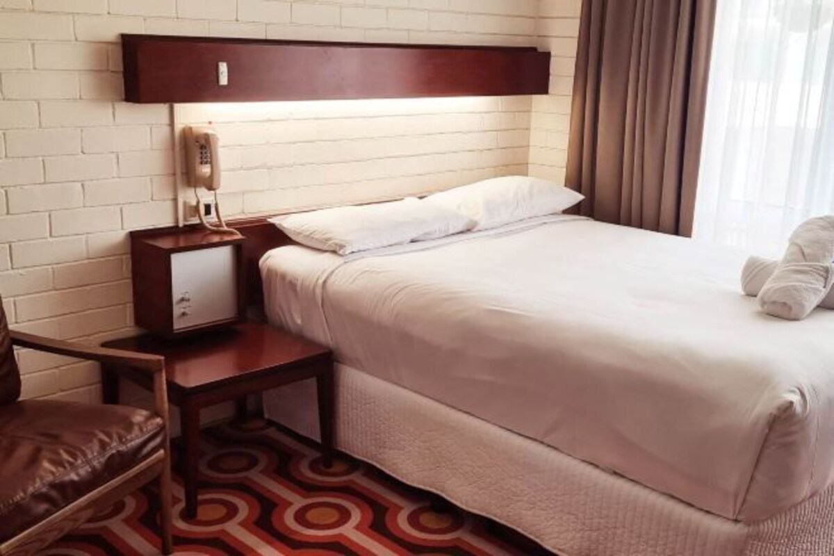 Een hotelkamer met een bed, een nachtkastje met een telefoon, een stoel en een kamerbreed tapijt met een patroon.