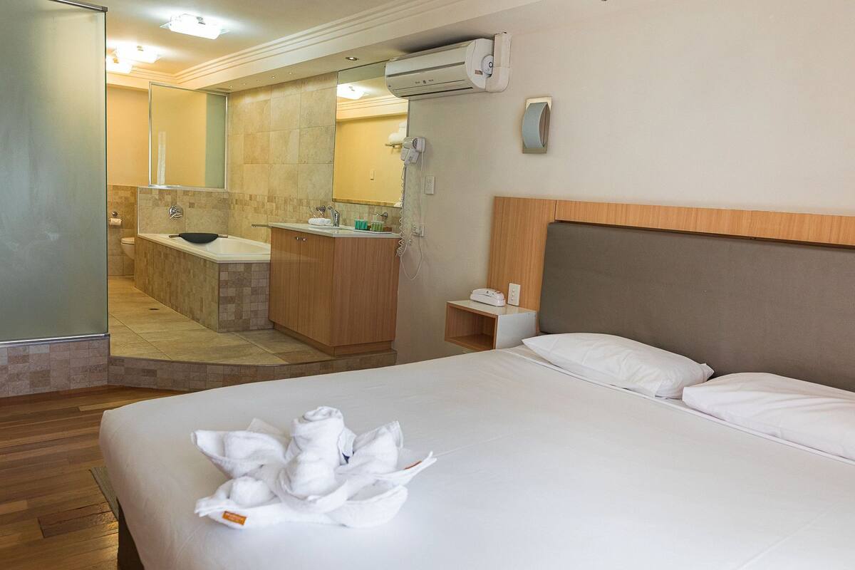 Ein Hotelzimmer mit Bett, einem Badezimmer mit Badewanne, einem hölzernen Kopfteil und einem Nachttisch mit Telefon und Wecker.