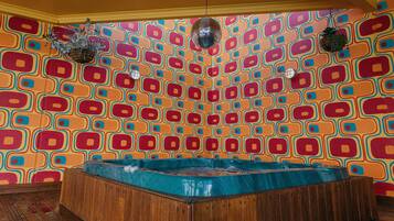 A room with a colorful geometric wall pattern, a wooden bathtub, and hanging plants.