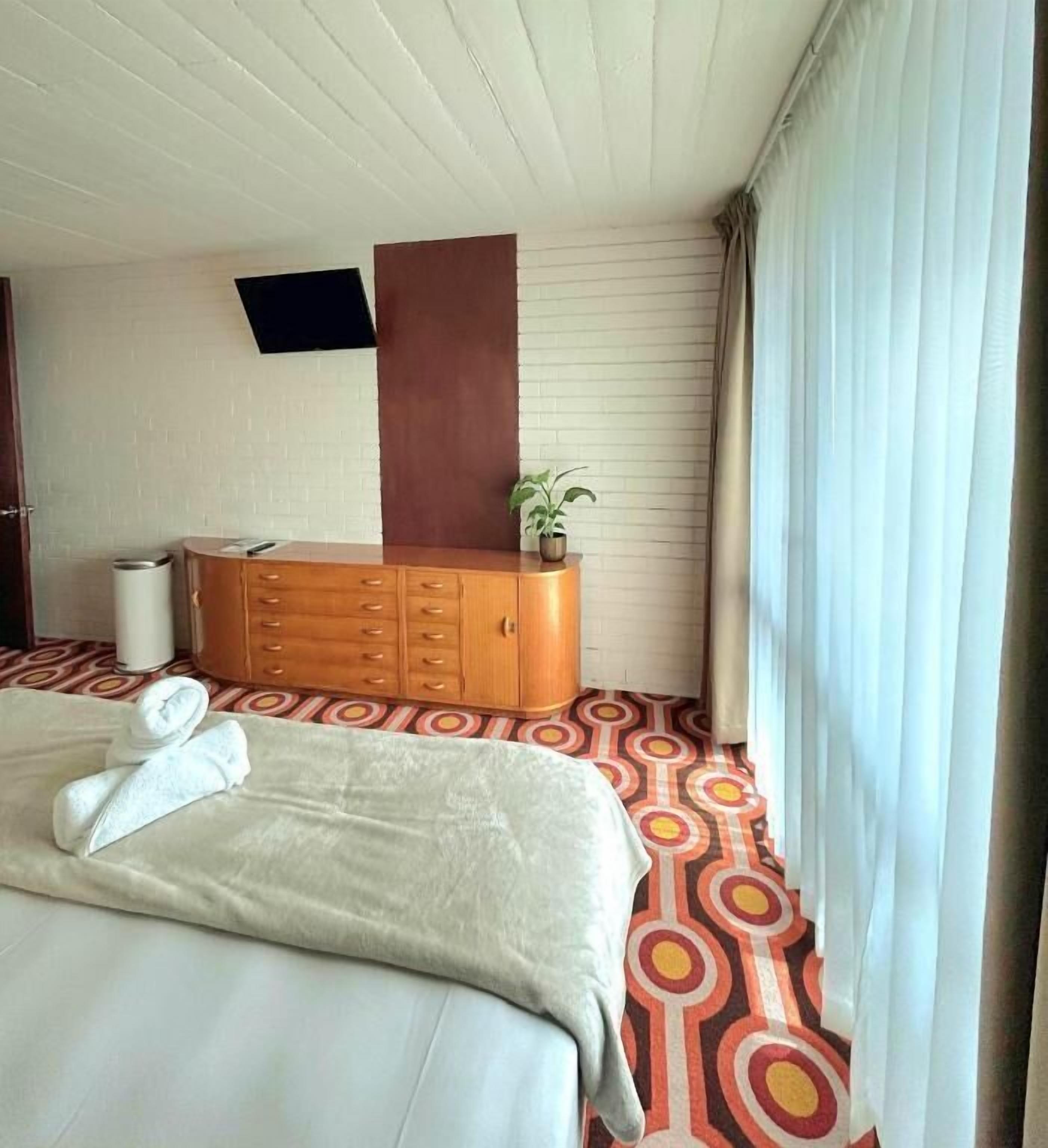 A hotel room with a bed, a wooden dresser, a potted plant, and a TV mounted on the wall.