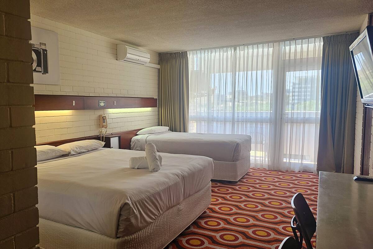 Ein Hotelzimmer mit zwei Betten, einer Ziegelwand, einer weißen Ziegelwand, einem Holz-Kopfteil, einem Telefon, einem Fenster mit Vorhängen und einem gemusterten Teppich.
