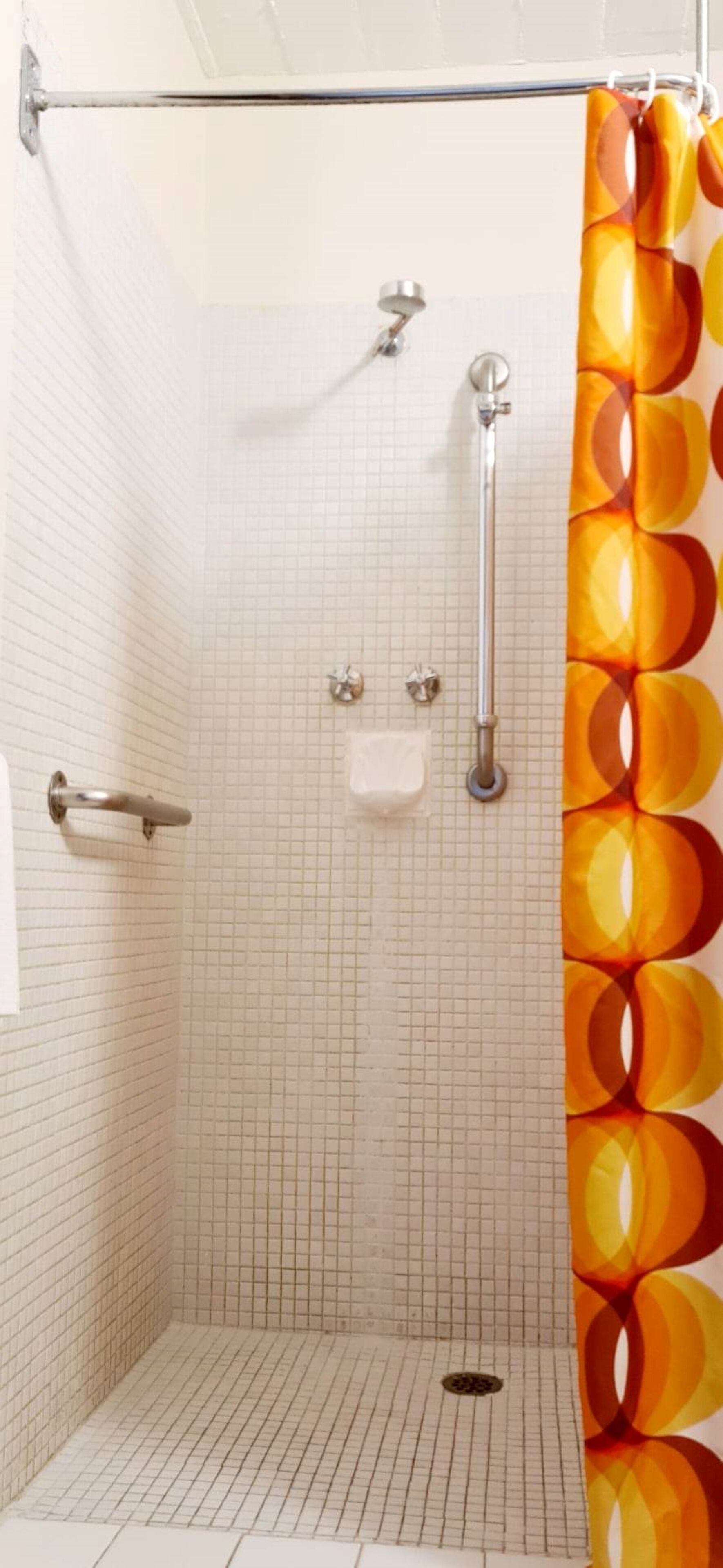 Une douche avec un mur recouvert de carreaux disposés en motif quadrillé, un pommeau de douche à main, un porte-serviettes et un rideau de douche orné d’un motif géométrique.