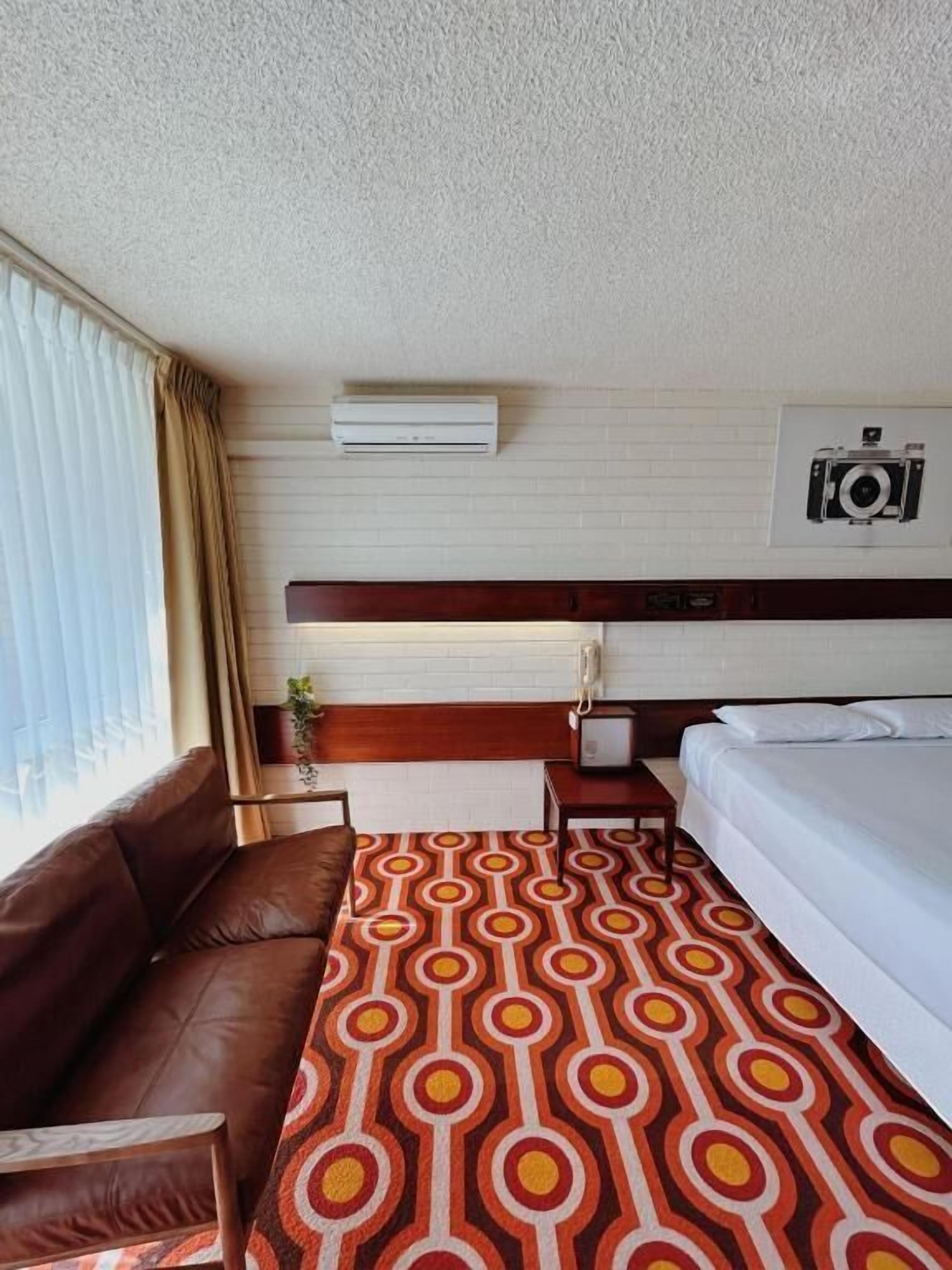 A hotel room with a brown leather sofa, a patterned carpet, a bed with white linens, an air conditioning unit, a mounted camera, and a wooden headboard.