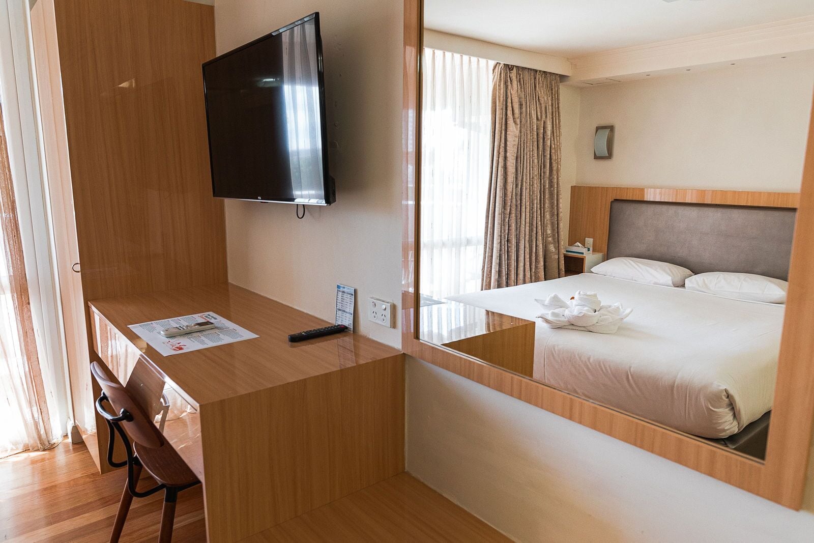 Habitación de hotel con escritorio de madera, televisor de pantalla plana, cama con sábanas blancas y un espejo que refleja la habitación.