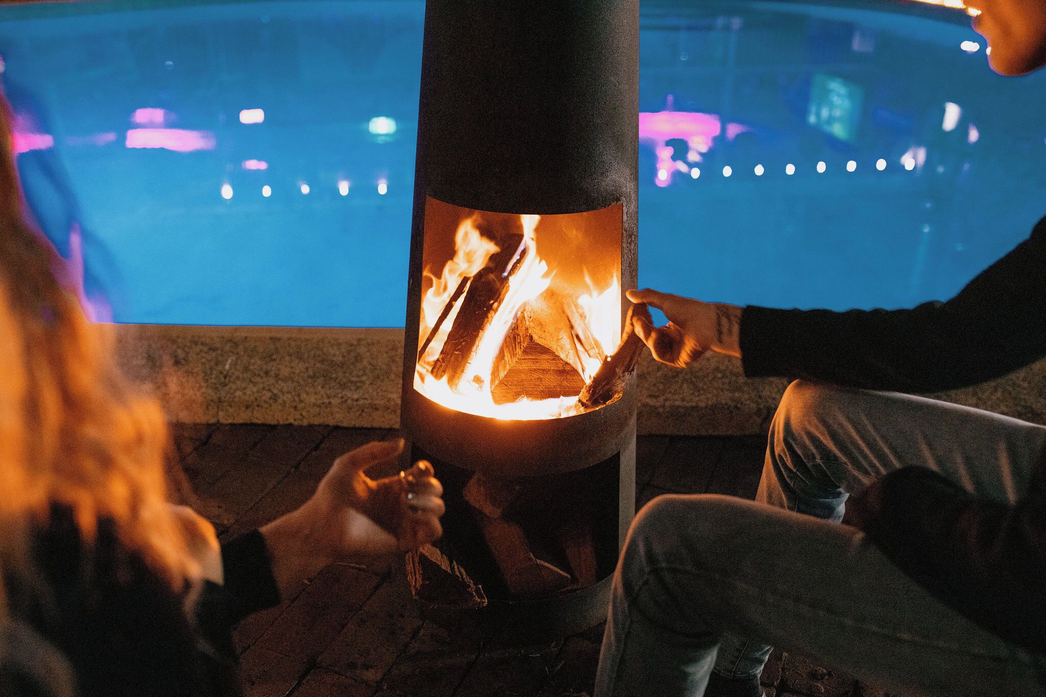 A person tending to a fire in a cylindrical outdoor heater near a pool.