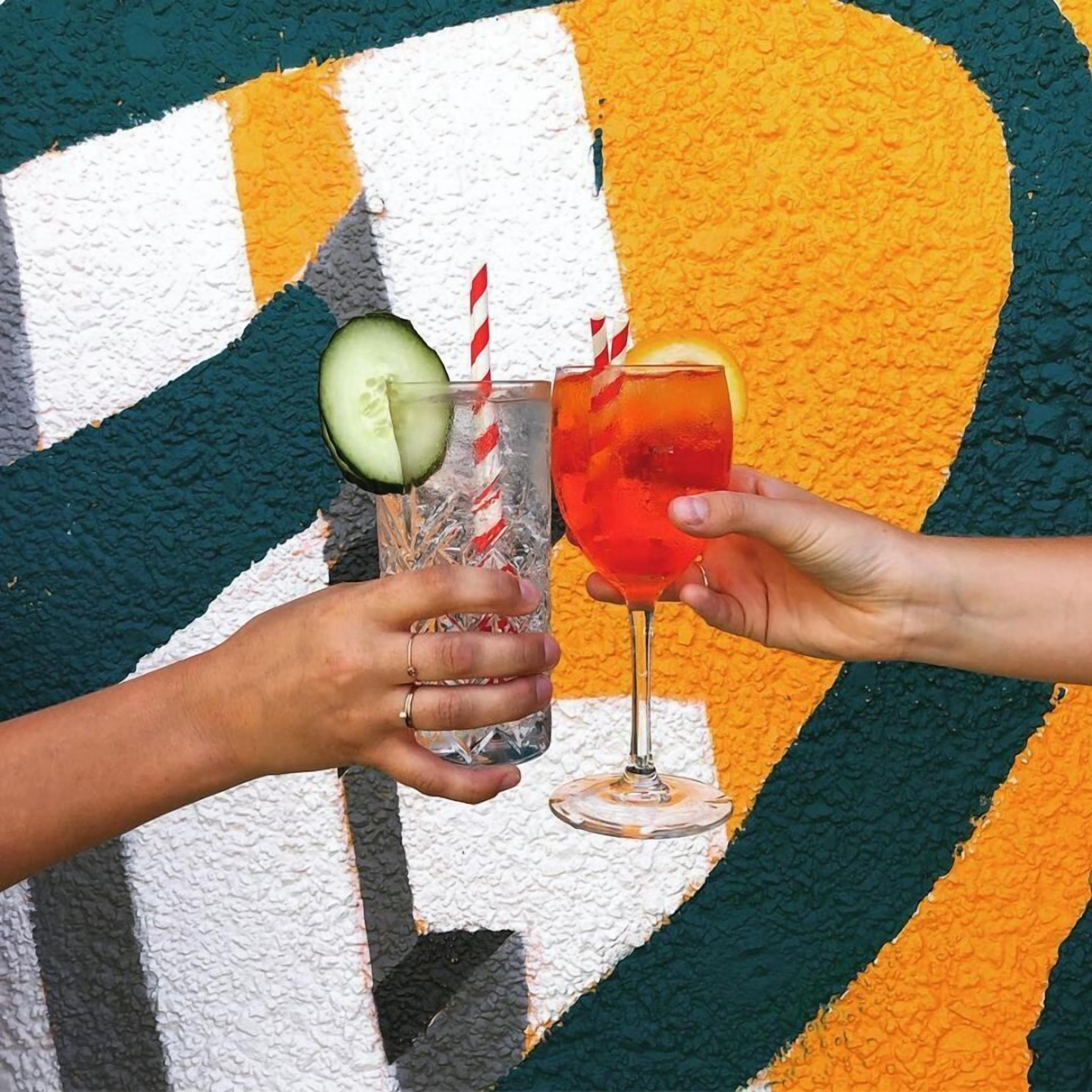 Two people holding glasses with drinks, one with a cucumber slice and a straw, the other with a red drink and a slice of orange.
