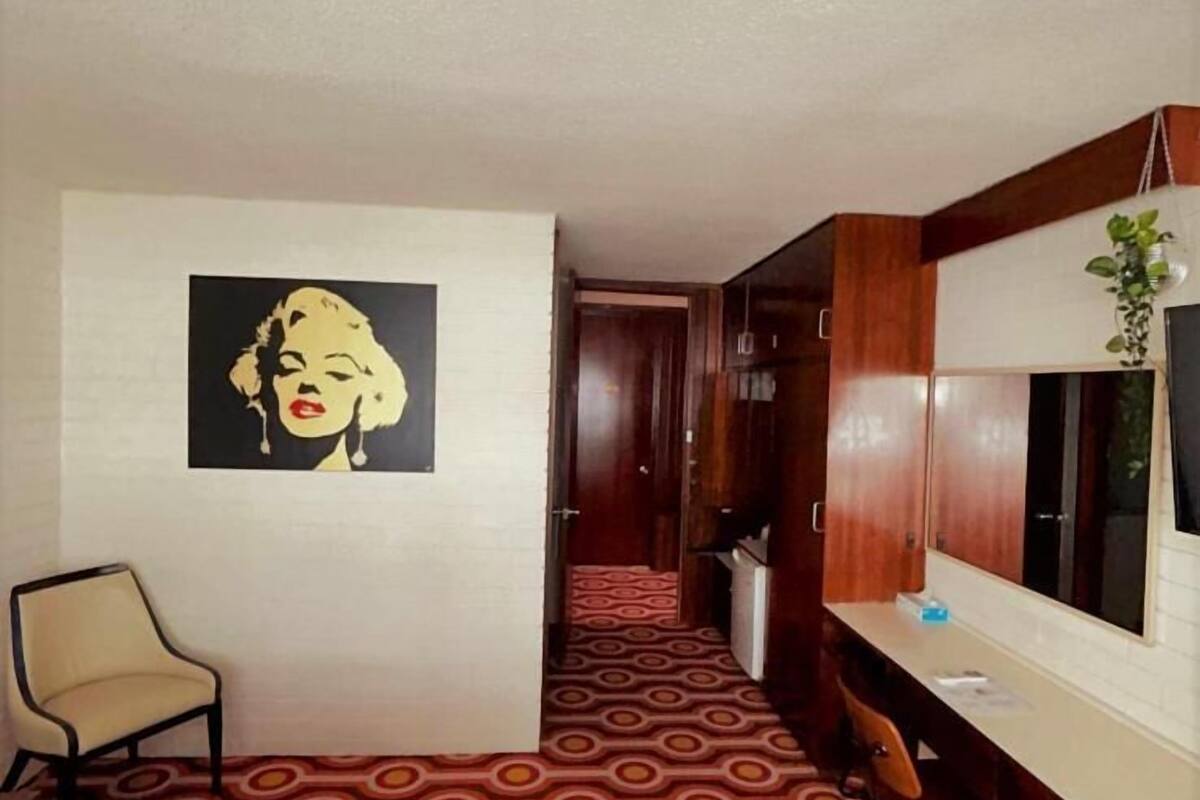 Ein Hotelzimmer mit Bett, Sessel, Schreibtisch, Spiegel, Kleiderschrank und einem Gemälde an der Wand.
