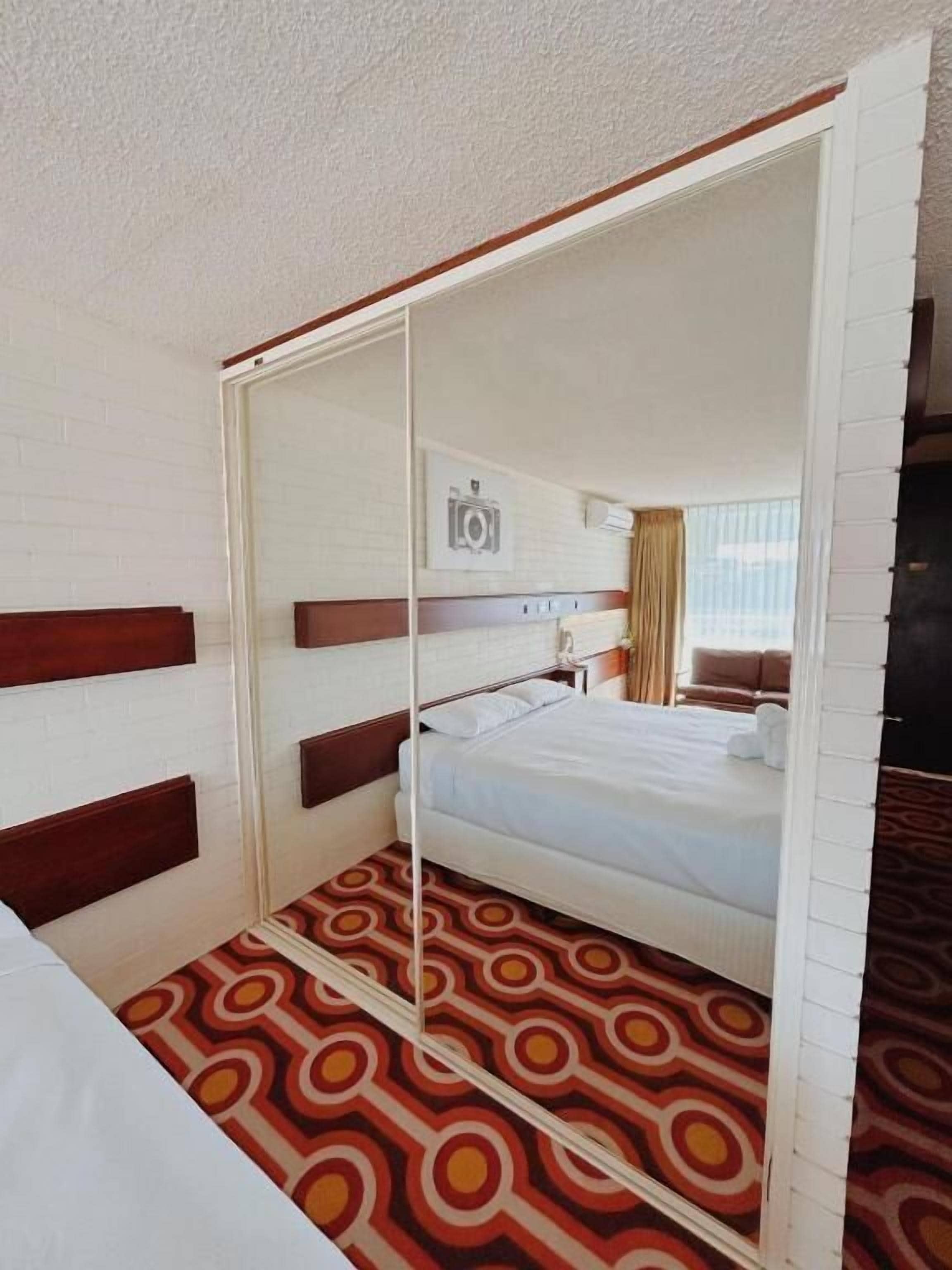 Habitación de hotel con cama, espejo, alfombra estampada y cámara de seguridad en la pared.