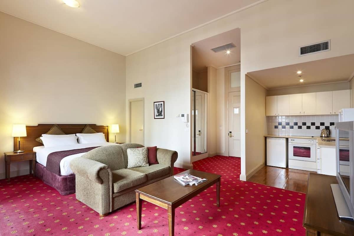 A hotel room with a bed, a sofa, a coffee table, a nightstand, a lamp, a red carpet, a kitchenette with a fridge, a stove, and a microwave.