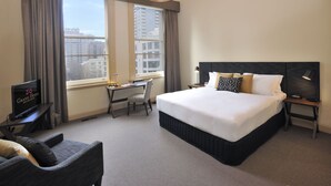 A hotel room with a large bed, a desk with a chair, a television, and a view of the city through the window. - Grand Hotel Melbourne (Docklands)