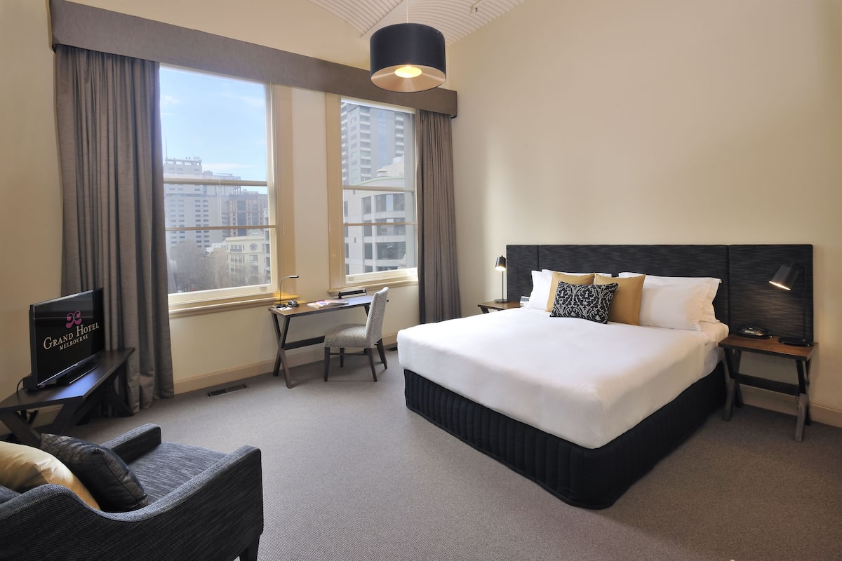 A hotel room with a large bed, a desk with a chair, a television, and a view of the city through the window.