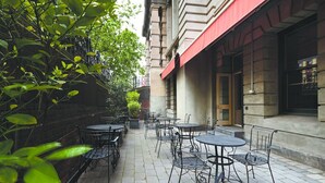 An outdoor patio with tables and chairs, surrounded by greenery and a building facade. - Grand Hotel Melbourne (Docklands)