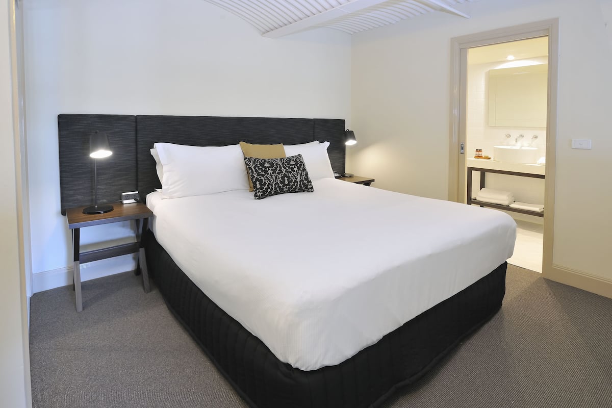 A neatly made bed with a dark headboard, a bedside table with a lamp, and a bathroom visible through an open door.