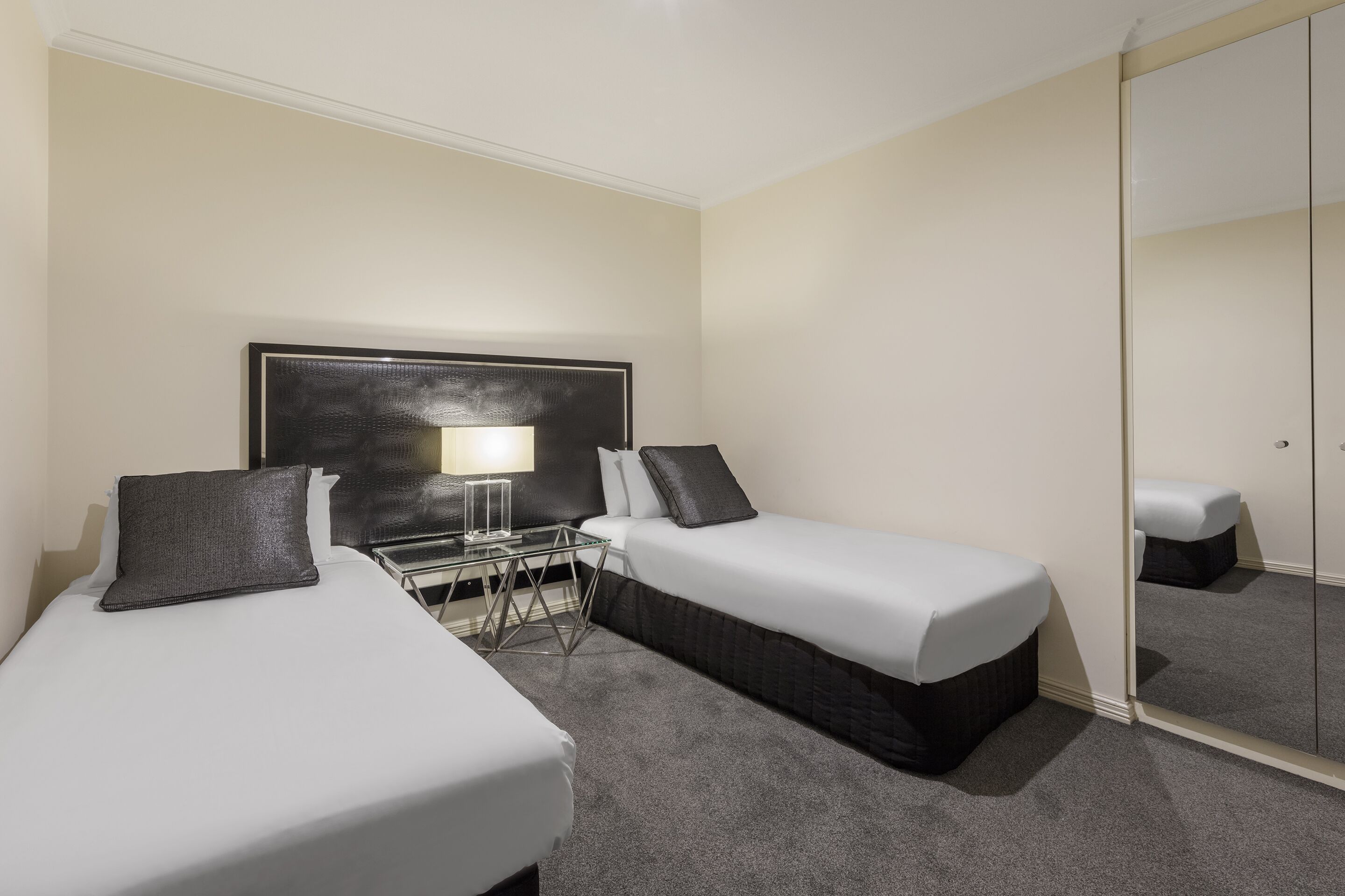 Habitación de hotel con dos camas individuales, una mesita de noche con lámpara, un espejo y piso alfombrado.