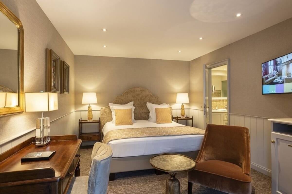 Een hotelkamer met een groot bed, twee nachtkastjes met lampen, een klein rond tafeltje, een bruine stoel, een televisie aan de muur en een spiegel.