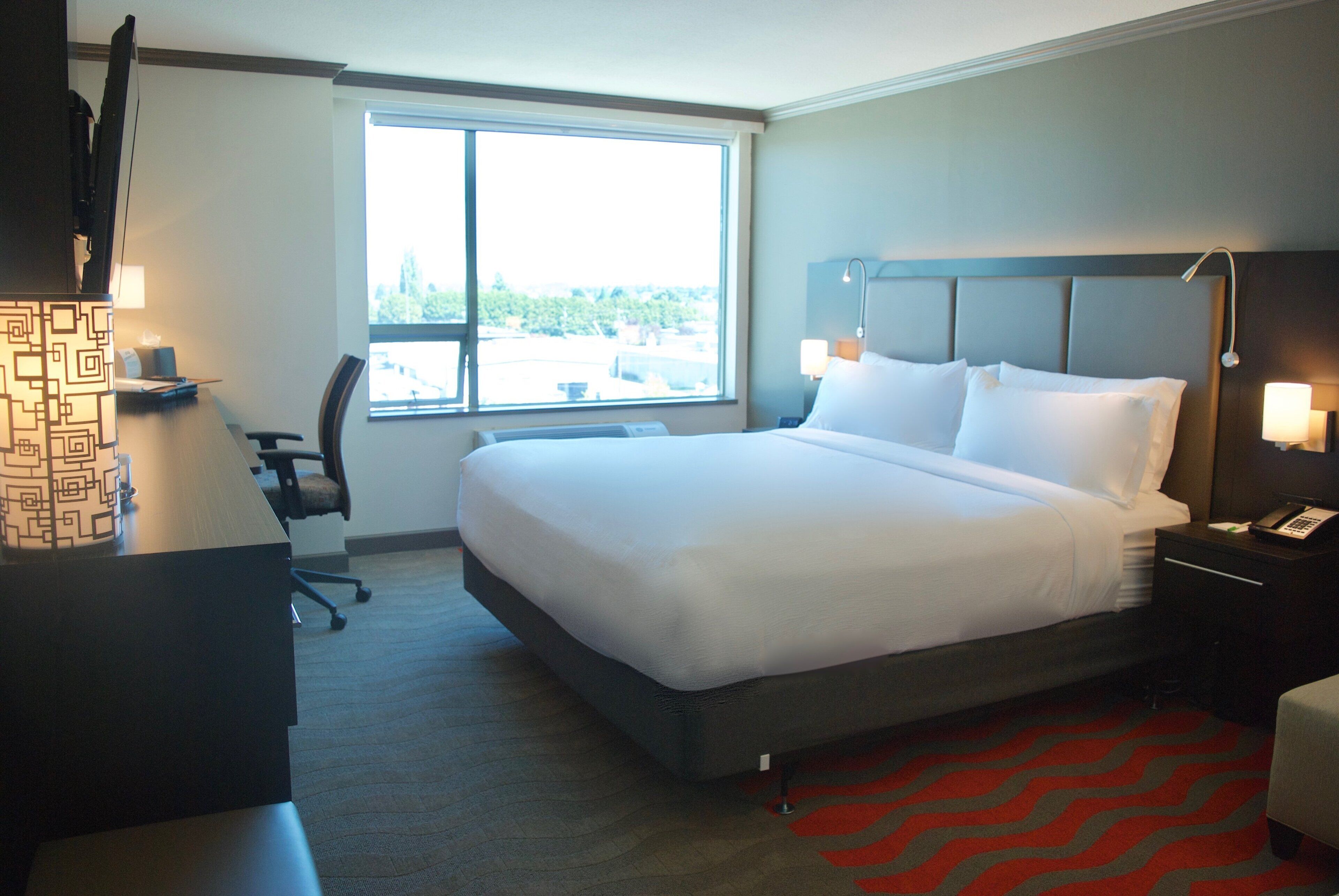 A hotel room with a large bed, a desk, a chair, a window, and a red carpet.