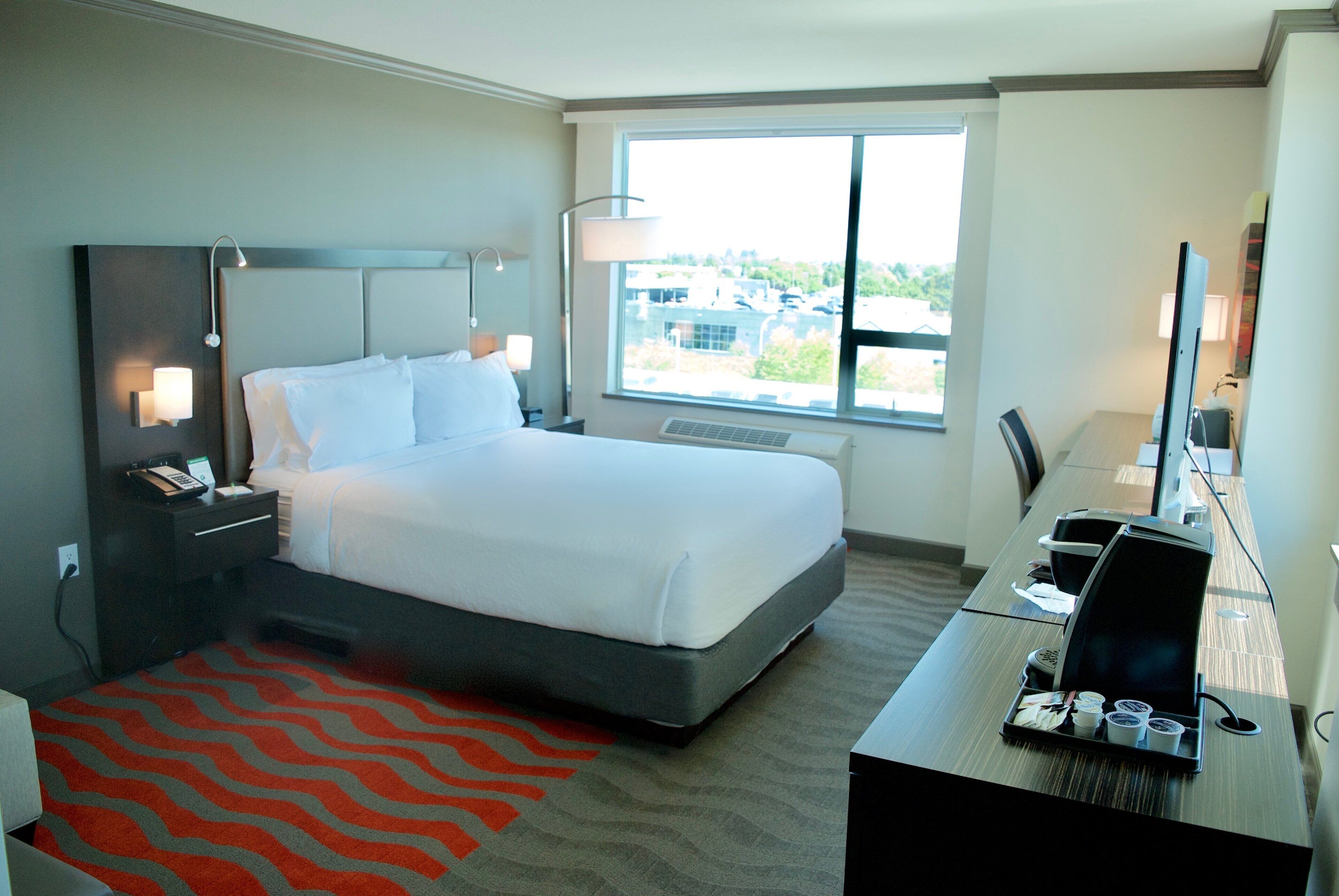 Quarto de hotel com cama, mesas de cabeceira, escrivaninha com computador, cadeira, janela com vista e tapete vermelho estampado.