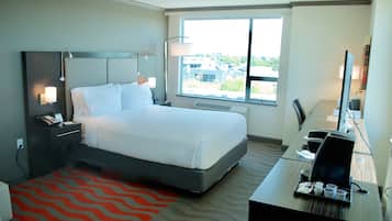A hotel room with a bed, bedside tables, a desk with a computer, a chair, a window with a view, and a red patterned rug.