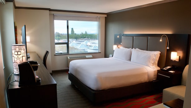 A hotel room with a large bed, a desk, a chair, a nightstand, and a window with a view of buildings and trees. - Holiday Inn Vancouver Airport- Richmond by IHG (Richmond)