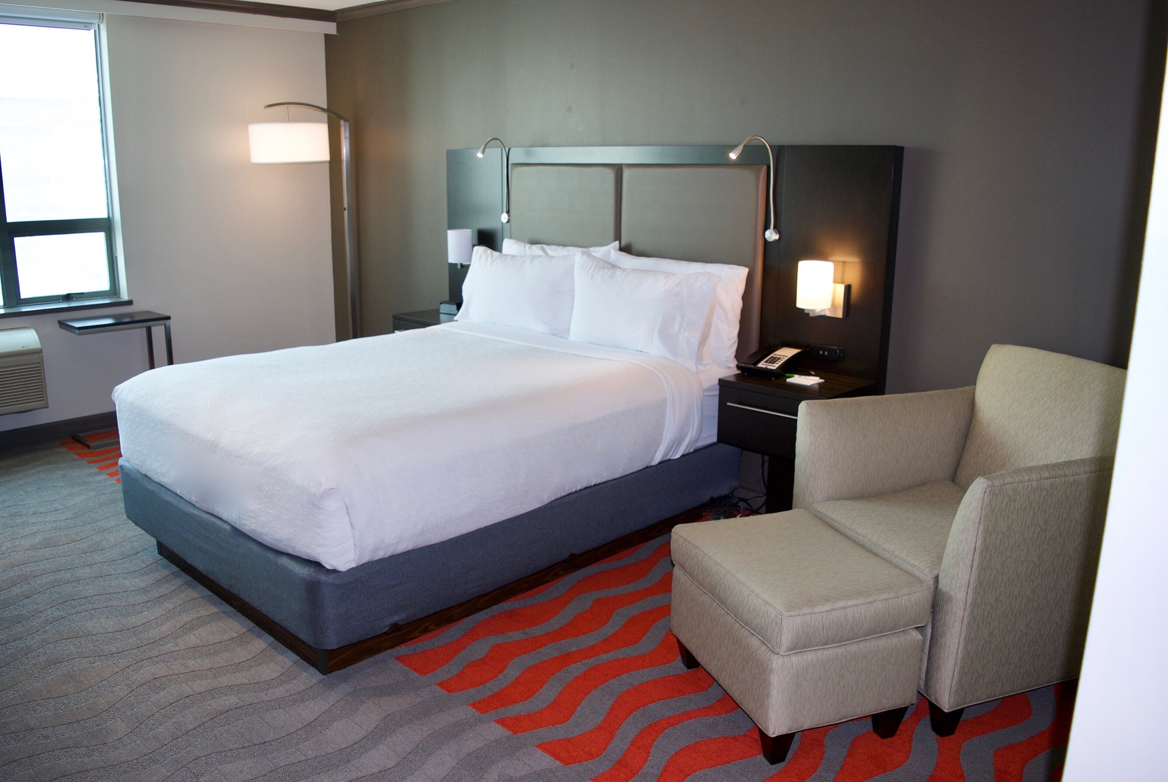 A hotel room with a large bed, a chair, a nightstand, a lamp, and a window.