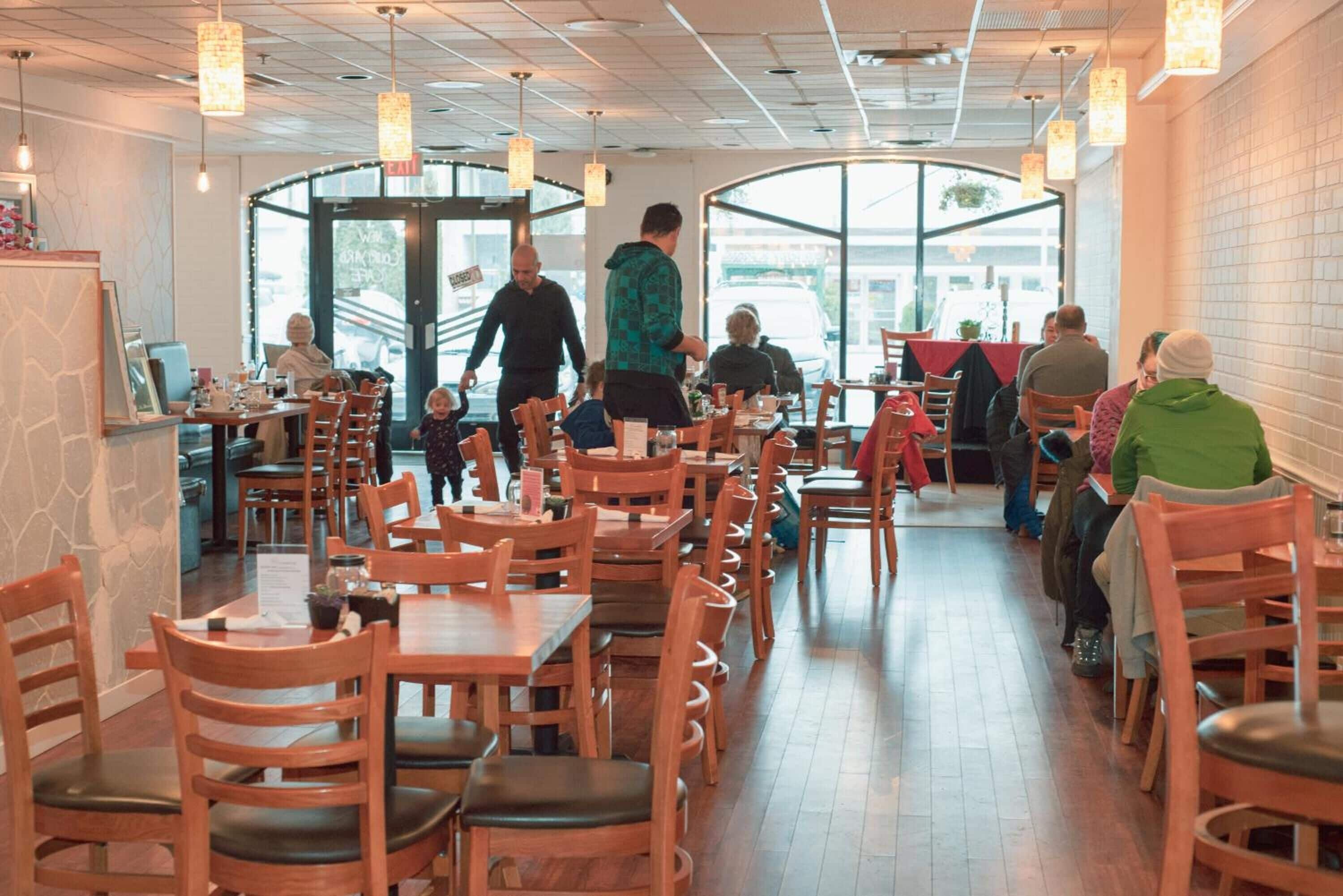 A restaurant with wooden tables and chairs, large windows, and hanging pendant lights.
