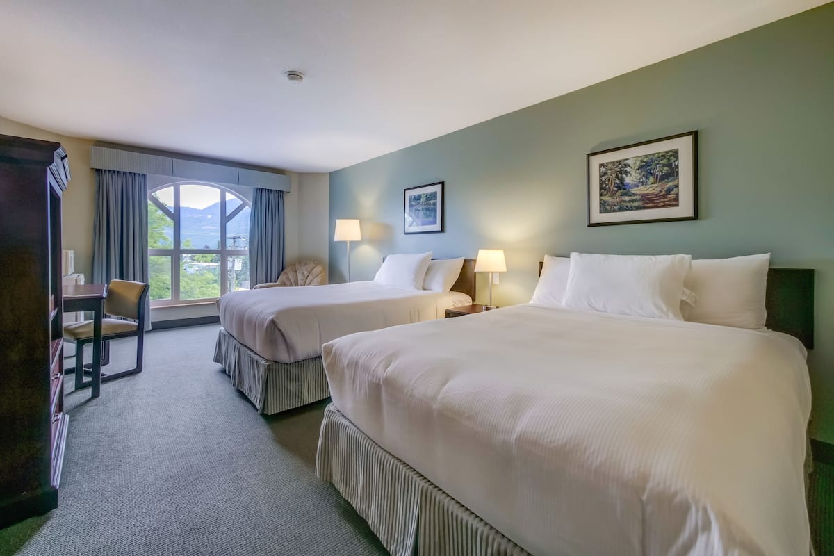 A hotel room with two beds, a chair, a desk, and a large window offering a view.