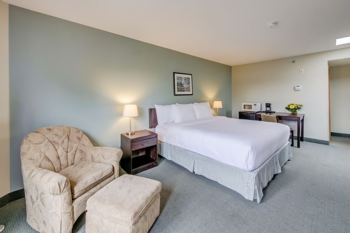 A hotel room with a bed, a chair, a small table with a lamp, a microwave, and a picture on the wall.