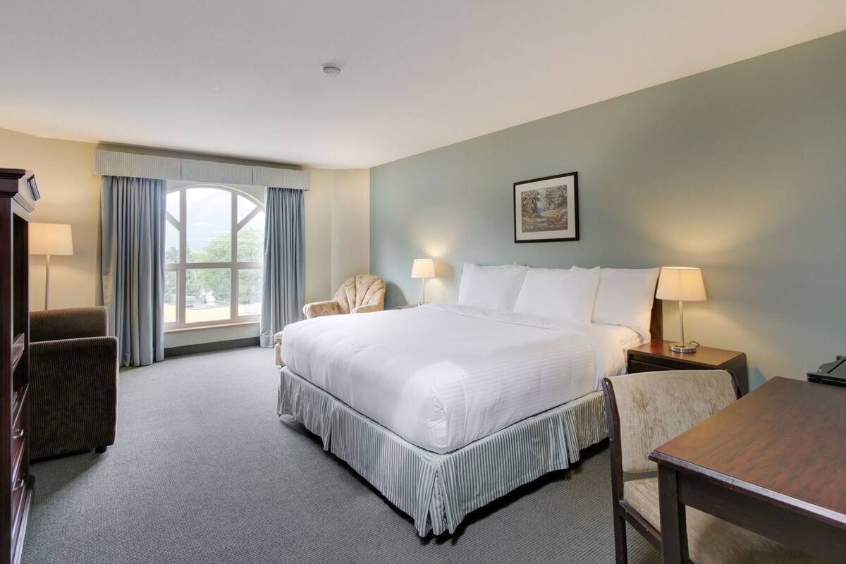 A hotel room with a bed, two chairs, a table, a window with curtains, a painting on the wall, and a lamp on the nightstand.