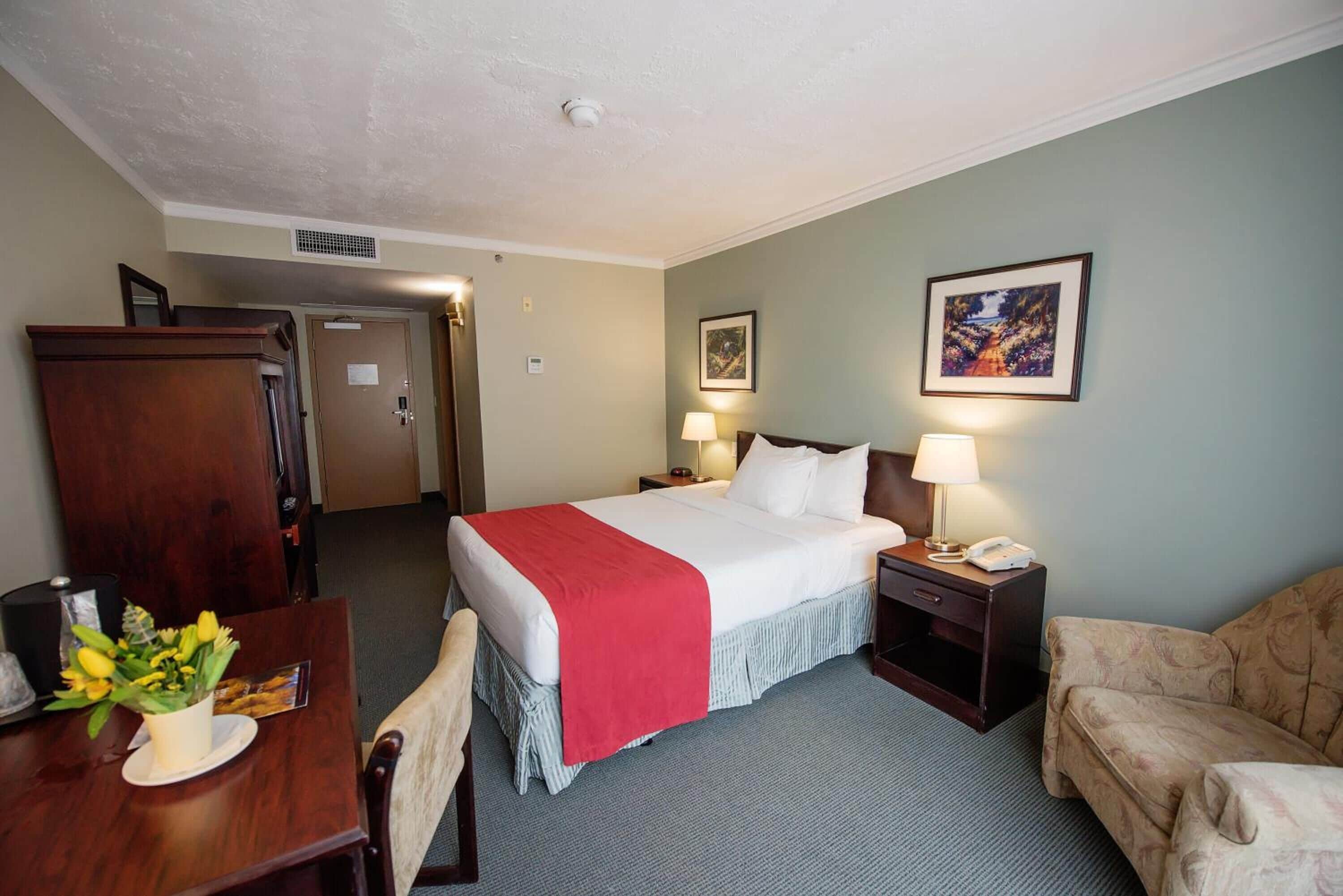 A hotel room with a bed, bedside tables, a desk with a chair, a couch, and a dining table with a vase of flowers.